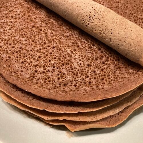 Sült injera palacsinták egy tányéron, egy tekercs injera látható a tetején.
